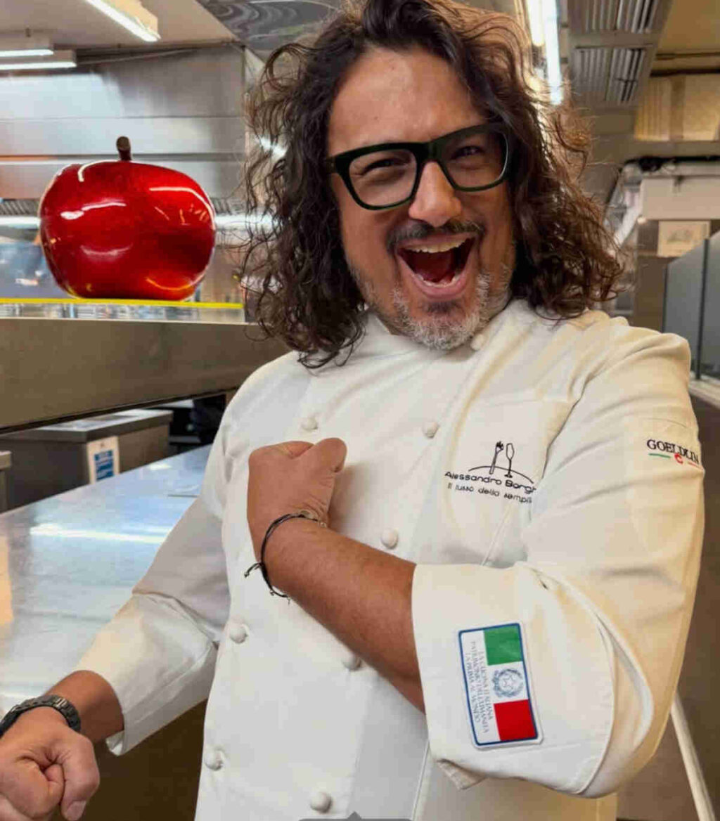 Alessandro Borghese nella cucina del suo ristorante che mostra orgoglioso lo scudetto della cucina italiana patrimonio dell'unesco