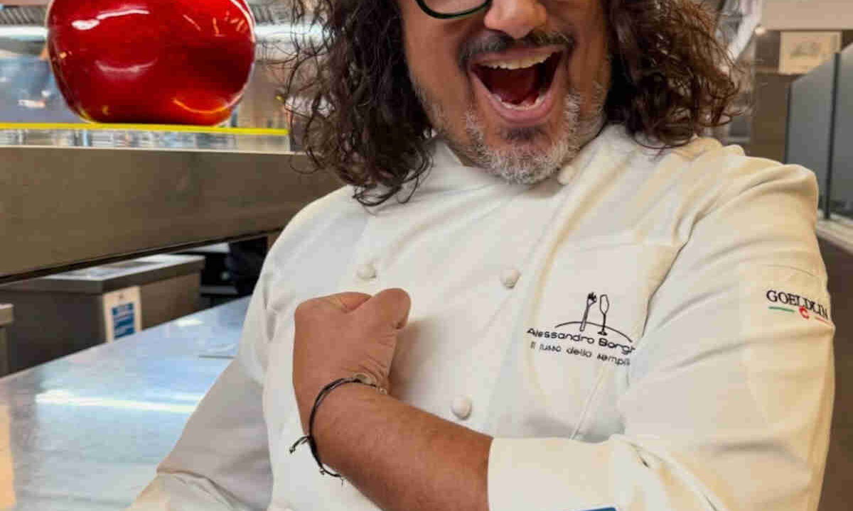 Alessandro Borghese nella cucina del suo ristorante che mostra orgoglioso lo scudetto della cucina italiana patrimonio dell'unesco