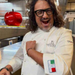 Alessandro Borghese nella cucina del suo ristorante che mostra orgoglioso lo scudetto della cucina italiana patrimonio dell'unesco