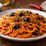 Piatto bianco su tavolo di legno con all'interno degli spaghetti alla sgualdrina secondo la ricetta di Ugo Tognazzi